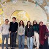 Six young adults stand smiling in front of a mural depicting symbols and names of various world religions.