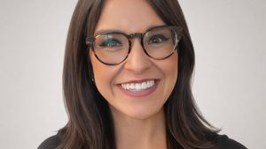 A woman with long dark hair and glasses smiles warmly against a plain light gray background.