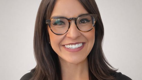 A woman with long dark hair and glasses smiles warmly against a plain light gray background.