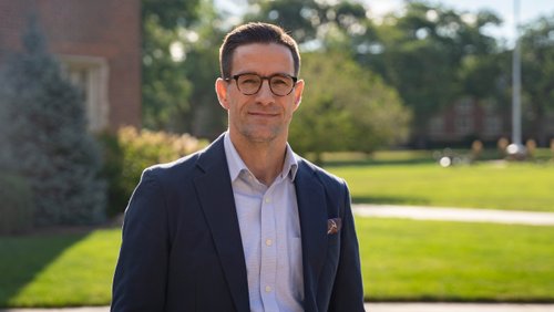 A man wearing glasses and a dark blazer smiles while standing outside on a sunny day with grass and trees behind him.