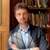 A JCU faculty member with curly brown hair and a beard, wearing a blazer, poses with crossed arms in front of a bookshelf.