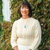 A woman with straight brown hair and red lipstick smiles wearing a cream cable-knit sweater and turquoise pendant.