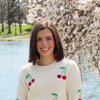 A smiling woman with straight brown hair wears a white sweater with red cherries and stands by a lake with blooming white.