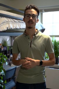 A young man with glasses and a goatee smiles while standing in a room with a bunk bed and a window behind him.