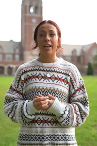 A smiling woman with red hair stands on a green lawn wearing a patterned sweater with a clock tower and brick building.