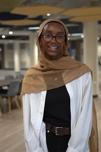 A smiling person wearing glasses, a tan headscarf, a white shirt, and a black top stands indoors with hands clasped.