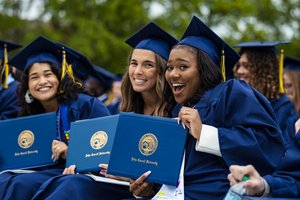 Three graduates in blue caps and gowns smile and hold up their John Carroll University diplomas outdoors.