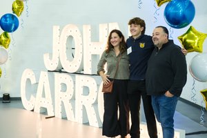 A student and his parents smile together in front of large illuminated John Carroll letters with blue and gold balloons.