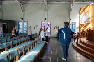 A man in a blue jacket gestures with raised hands toward a small group standing near stained glass windows in a church.
