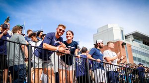 A group of young people stand behind a black railing in a stadium, some smiling and clapping under a blue sky.