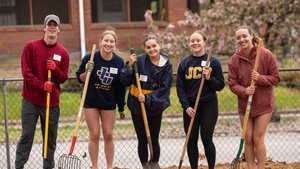 Five young people stand outside holding gardening tools and smiling in front of a chain-link fence and a brick building.
