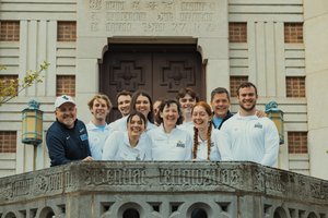 A group of people wearing white JDOS shirts smile and pose together on a stone balcony outside Grasselli Library.