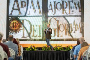 A man stands on a stage speaking into a microphone in front of an audience and a large screen reading Ad Majorem Dei Gloriam.
