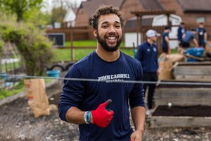 A smiling young man wearing a John Carroll Football shirt and red gloves gives a thumbs-up in a garden area.