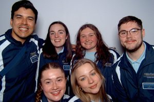 Six young adults wearing matching John Carroll University tour guide jackets smile closely together against a plain wall.