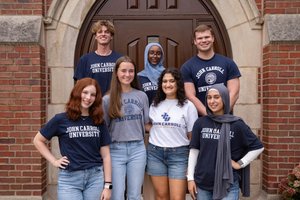 Seven diverse young adults wearing John Carroll University shirts smile and pose in front of a large wooden door in a stone archway.