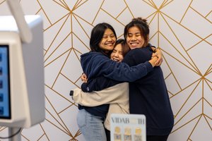 Three young women smile with their eyes closed as they hug each other in front of a geometric photo booth backdrop.
