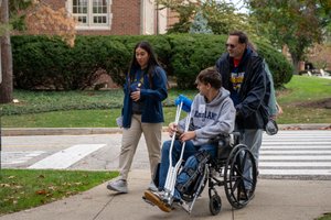 A man in a wheelchair with a leg brace and crutches is pushed by another man while a woman walks beside them talking.