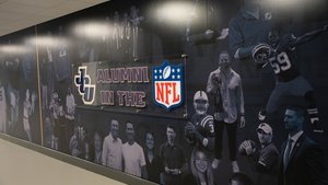 A wall mural displays black and white images of people and athletes with a sign reading JCU Alumni in the NFL.