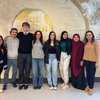 Eight people stand side by side smiling in front of a wall mural featuring a globe and religious symbols.