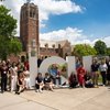 A group of people pose around large white letters spelling JCU on a sunny day with a tall brick clock tower and trees behind.