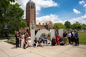A group of people pose around large white letters spelling JCU on a sunny day with a tall brick clock tower and trees behind.