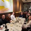 Six people sit around a table in a restaurant with wine glasses, menus, and bread baskets under a large abstract yellow.