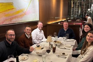 Six people sit around a table in a restaurant with wine glasses, menus, and bread baskets under a large abstract yellow.