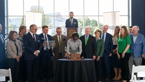 A group of people stand around a table as a woman places a brick on a wooden base during a ceremony in front of a large.