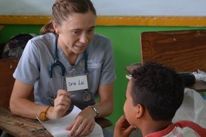 A woman wearing scrubs and a stethoscope talks to a boy sitting at a desk while holding a paper.