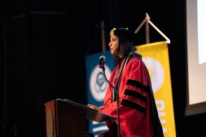A speaker in red academic regalia, with an Alpha Sigma Nu stole, addresses an audience from a wooden podium at a university