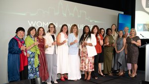 Thirteen women stand side by side on a stage holding glasses and smiling in front of a screen displaying "Women of Influence.