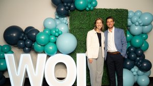 A man and a woman stand smiling in front of a green hedge wall decorated with teal and dark blue balloons and large.