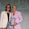 Two women smiling and standing close together, one holding a decorative glass award, with a city skyline drawing behind them.