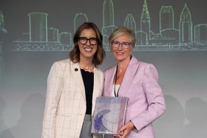 Two women smiling and standing close together, one holding a decorative glass award, with a city skyline drawing behind them.