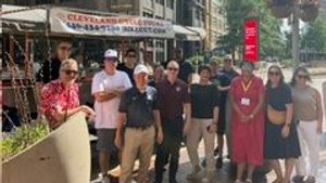 A group of eleven people stand on a city sidewalk near a street vendor and tall buildings under a partly cloudy sky.