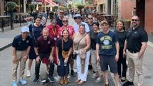 A group of people posing and smiling together on a city street lined with buildings and outdoor seating under a partly.