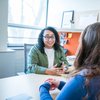 A person with curly hair and glasses talks animatedly across a desk to another person with long hair.