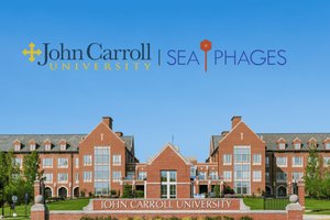 A large brick university building with many windows stands behind a sign reading John Carroll University under a clear blue.