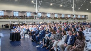 A large audience seated and standing inside a gymnasium attentively watching an event near a table with a black cloth.