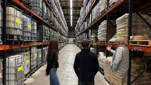 Three people stand in a warehouse aisle lined with tall shelves holding large containers and bags.