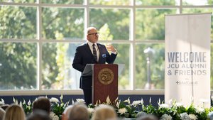 A man in a suit speaks at a podium with a university seal in front of an audience and a banner reading welcome JCU alumni.