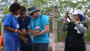 Three people in scrubs and one person in a blue shirt and hat review clipboards while another woman films with a handheld.