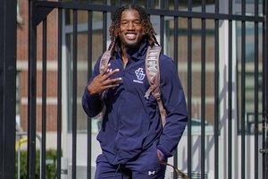 A man wearing a navy blue football tracksuit and backpack smiles while holding up four fingers in front of a black metal.