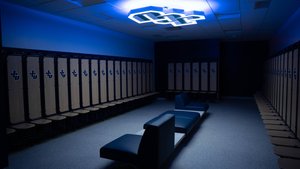 A dimly lit locker room with rows of wooden lockers featuring a logo and a modern blue bench in the center beneath a glowing.