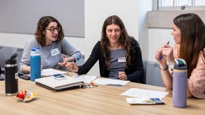 Three women sit at a table with papers and water bottles, engaged in a lively discussion in a bright room.