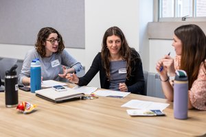 Three women sit at a table with papers and water bottles, engaged in a lively discussion in a bright room.