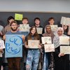 A diverse group of eleven people stand in front of a blackboard holding colorful handwritten signs with artistic designs and