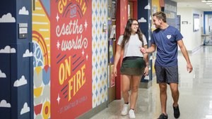 A young woman and man walk and talk smiling in a brightly decorated hallway with a large red mural reading go forth and set.