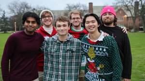 Six young men stand close together on a grassy area, smiling at the camera with trees and buildings behind them.